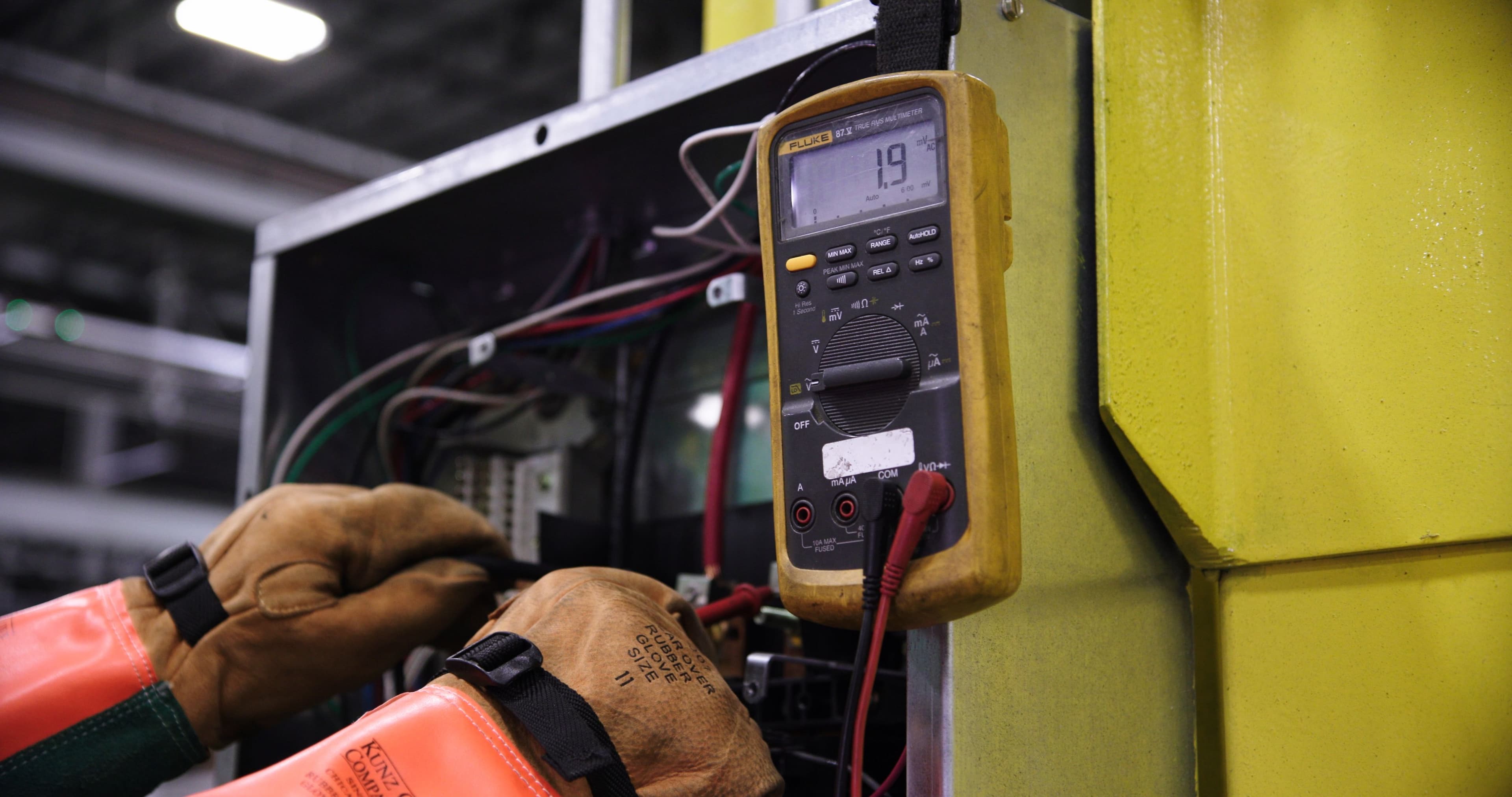 Gloved hands checking terminations with a multimeter inside an open control panel